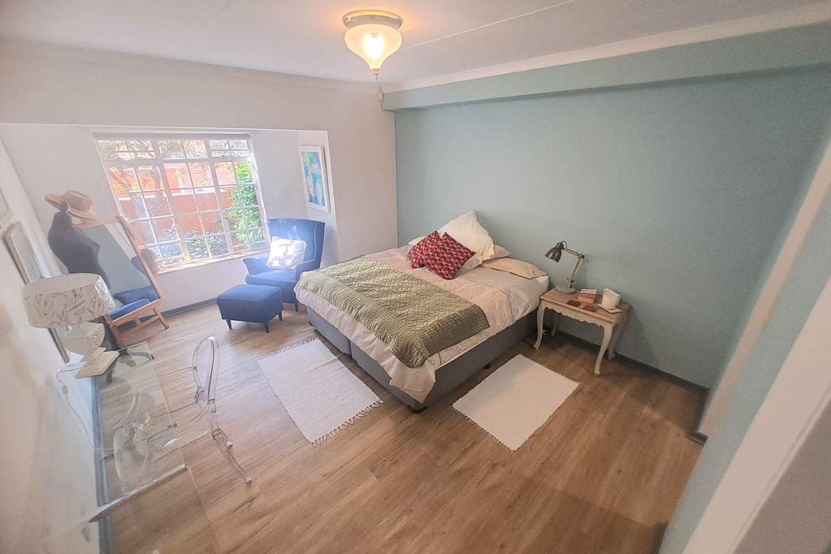 A well-lit bedroom featuring light-toned laminate flooring, a soft blue accent wall, and a large window providing natural light.