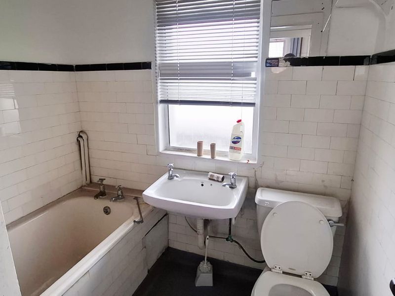 A dated bathroom featuring a standard bathtub, wall-mounted basin, and toilet with classic white ceramic wall tiling and black accent trim.