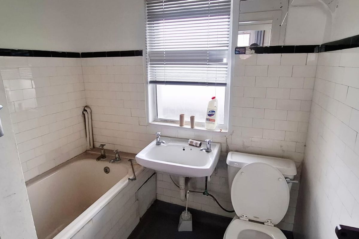 A dated bathroom featuring a standard bathtub, wall-mounted basin, and toilet with classic white ceramic wall tiling and black accent trim.