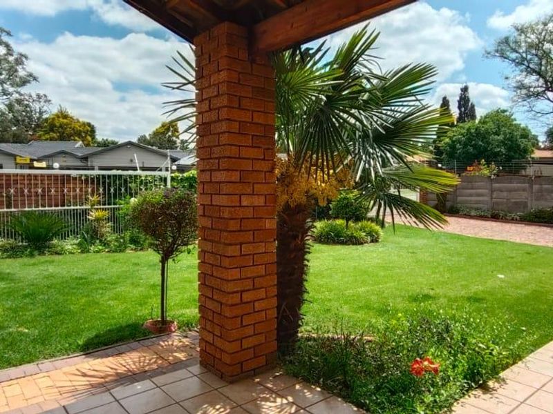 A covered patio area featuring brick pillars and tiled flooring, overlooking a well-maintained garden and paved driveway.