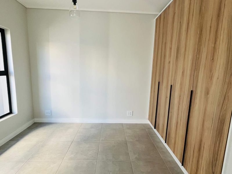 A bright, modern bedroom featuring neutral tiled flooring, clean white walls, and floor-to-ceiling wood-finish built-in cupboards with sleek black handles.