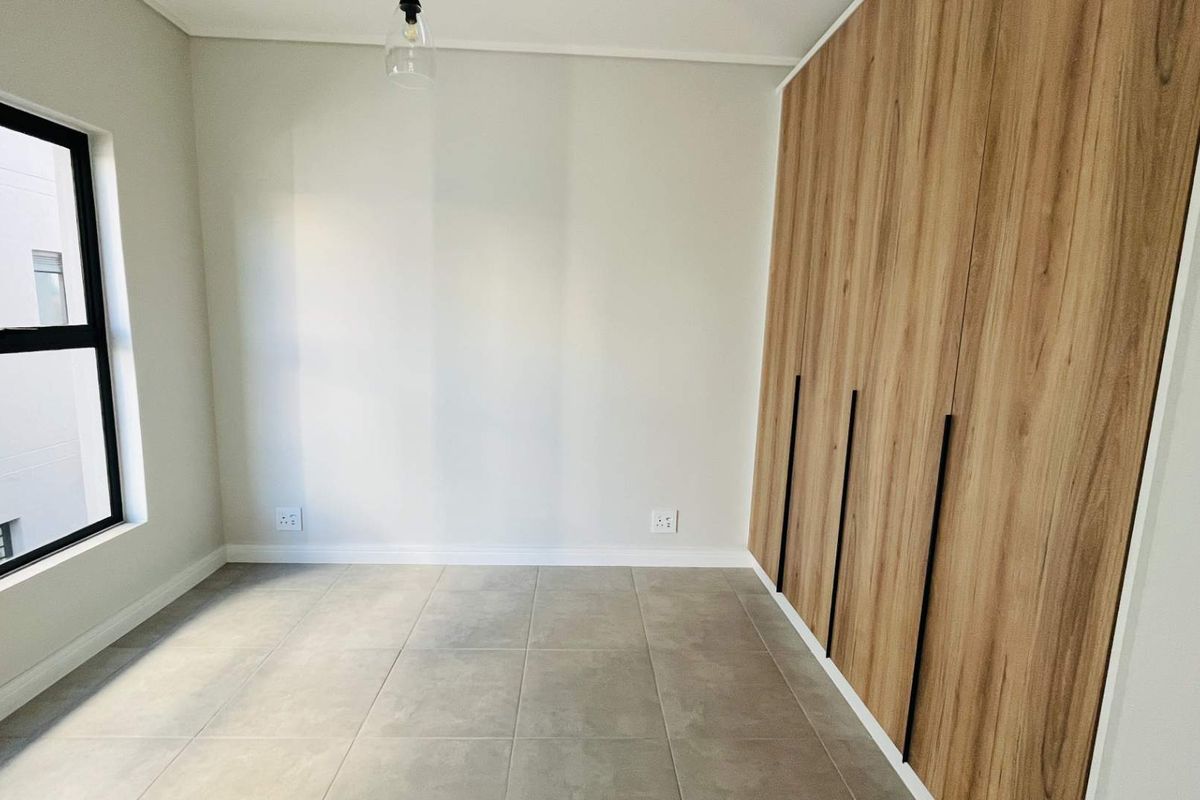 A bright, modern bedroom featuring neutral tiled flooring, clean white walls, and floor-to-ceiling wood-finish built-in cupboards with sleek black handles.