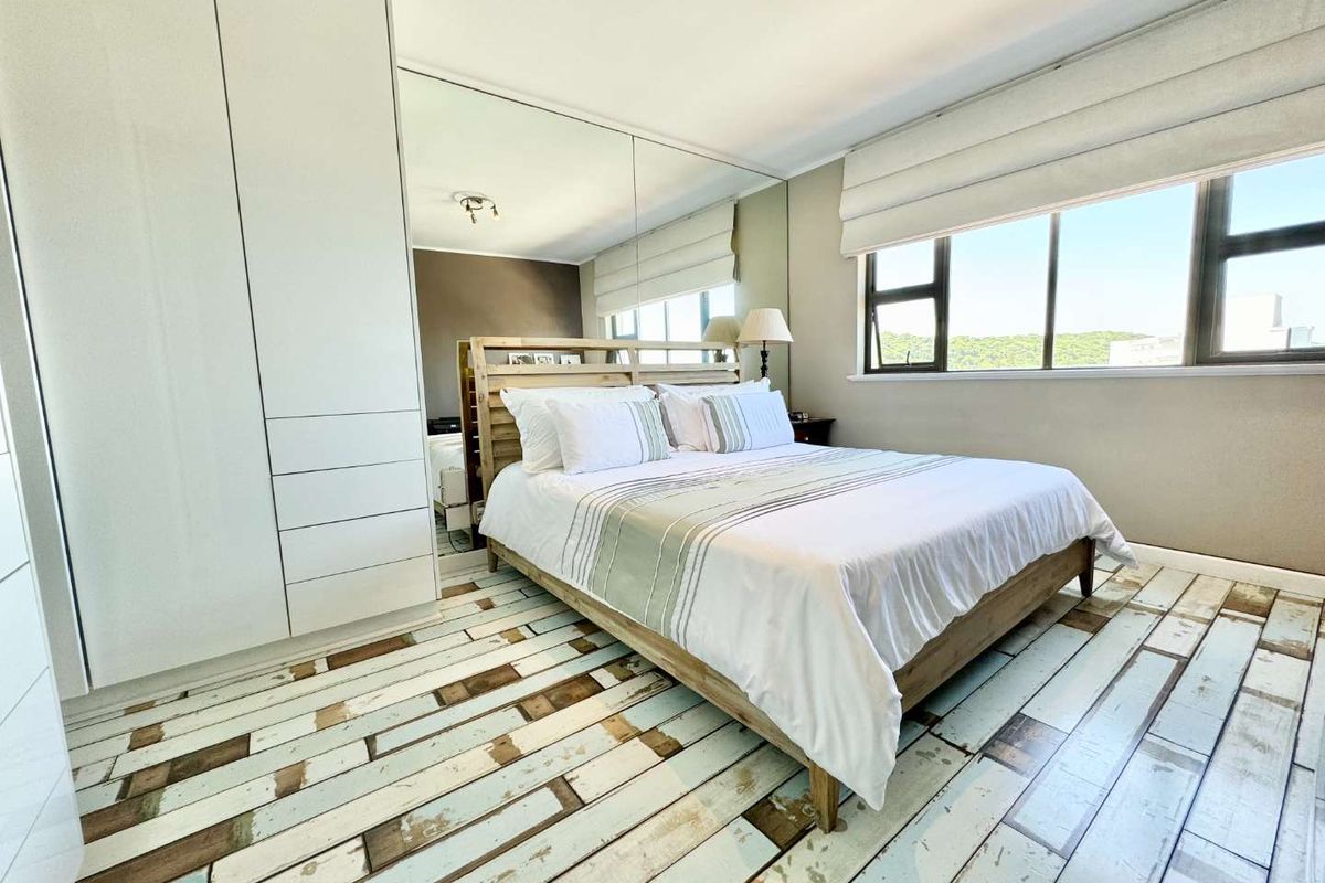 A bright bedroom featuring built-in floor-to-ceiling wardrobes, a large mirrored wall panel, and a window with fitted roman blinds.