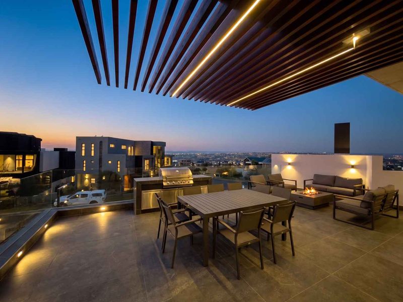 A modern, covered outdoor patio featuring a slatted timber pergola with integrated lighting, a built-in stainless steel braai, and panoramic estate views.