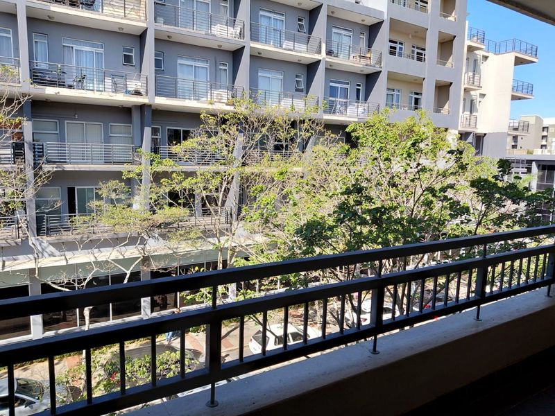 A spacious apartment balcony featuring a metal balustrade and views of the surrounding building complex and mature trees.