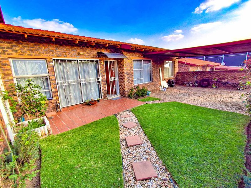 Front view of a face-brick cluster home featuring a paved driveway, covered patio entrance, and a well-maintained garden with a stone pathway.