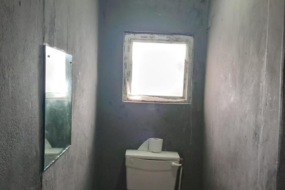 A small, unfinished bathroom featuring exposed cement walls, an exposed timber ceiling, and a basic toilet installation, requiring significant renovation and finishing work.