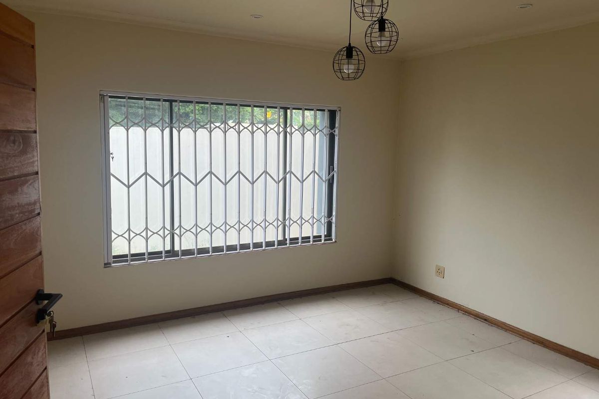 A bright, empty bedroom featuring neutral tiled flooring, a large window with security bars, and modern pendant lighting.