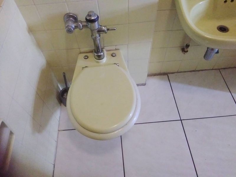 A dated bathroom featuring a wall-mounted toilet with a flush valve and a wall-mounted basin, finished with ceramic wall and floor tiles.