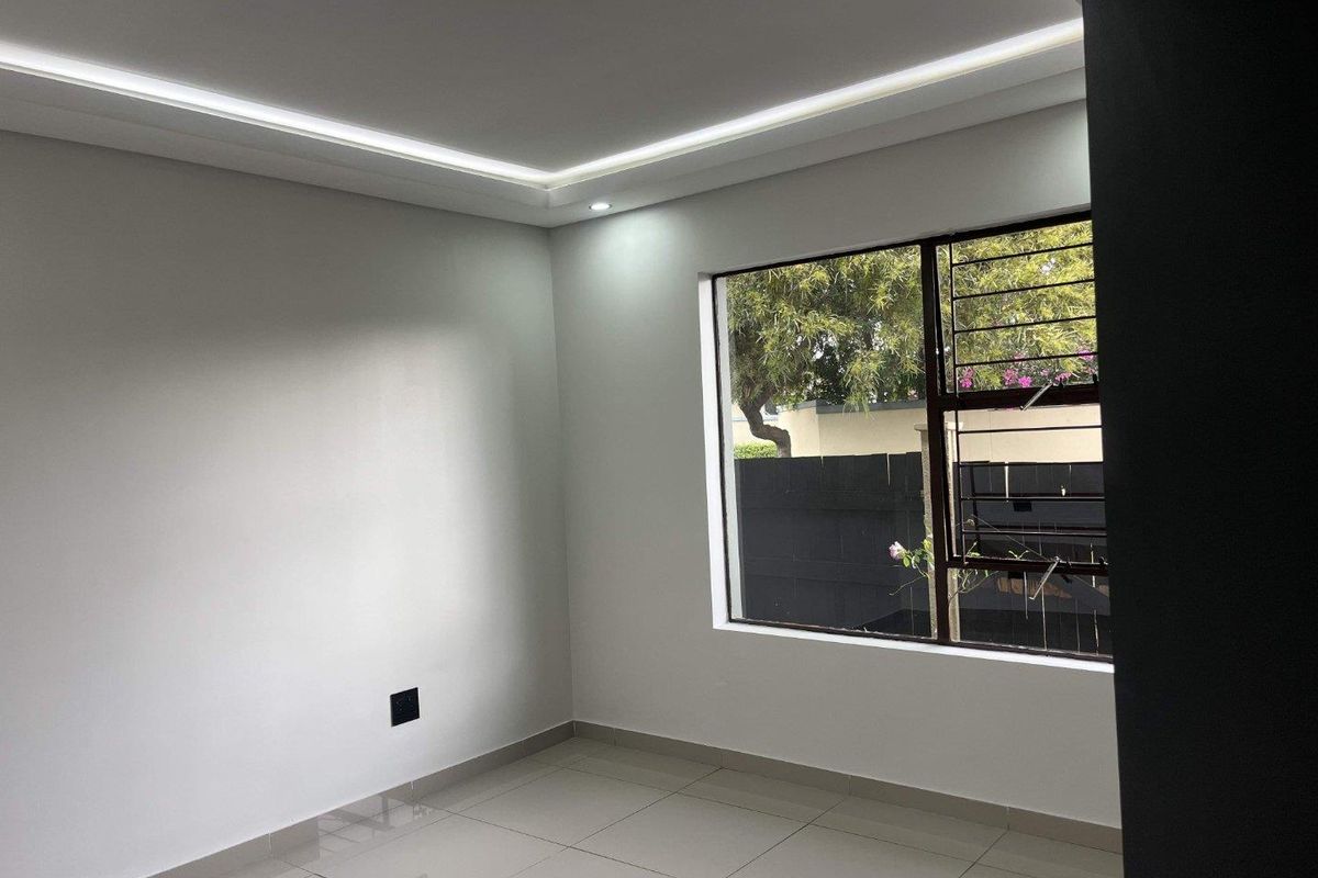 A bright, empty bedroom featuring modern recessed ceiling lighting, large windows with security bars, and neutral tiled flooring.
