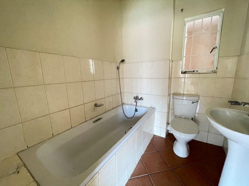 A dated bathroom featuring a bathtub with shower mixer, pedestal basin, and toilet, finished with ceramic wall tiles and terracotta floor tiling.