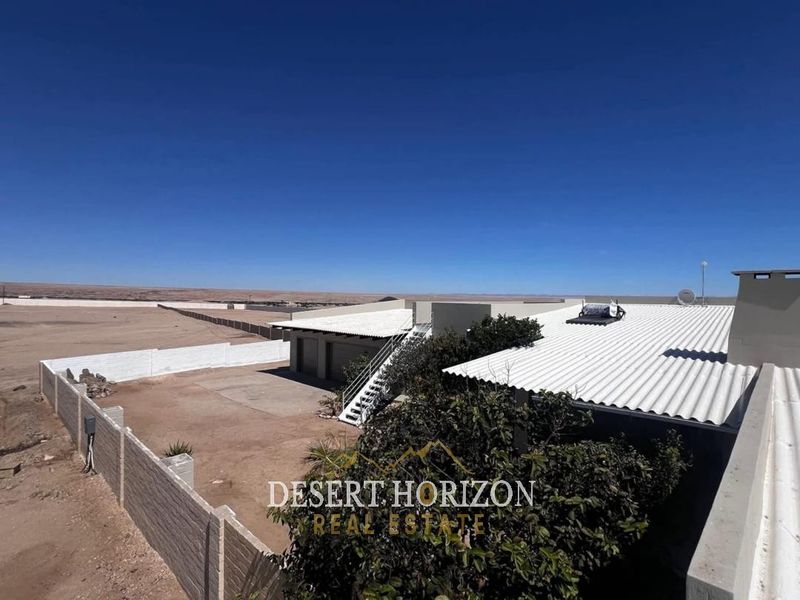 Aerial view of the property showing the corrugated metal roof, solar water heating system, and surrounding perimeter boundary walls in a desert landscape.