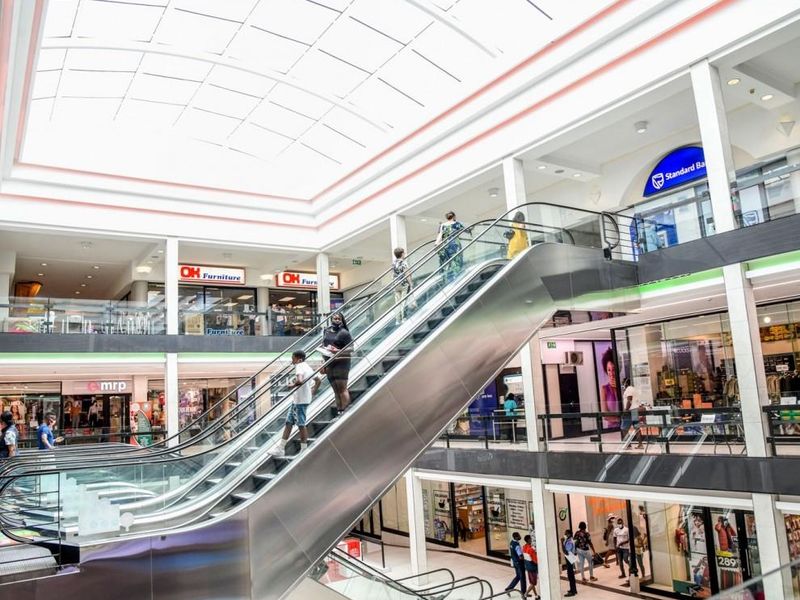 Interior view of a multi-level retail shopping center featuring a central escalator, glass railings, and a large overhead skylight.