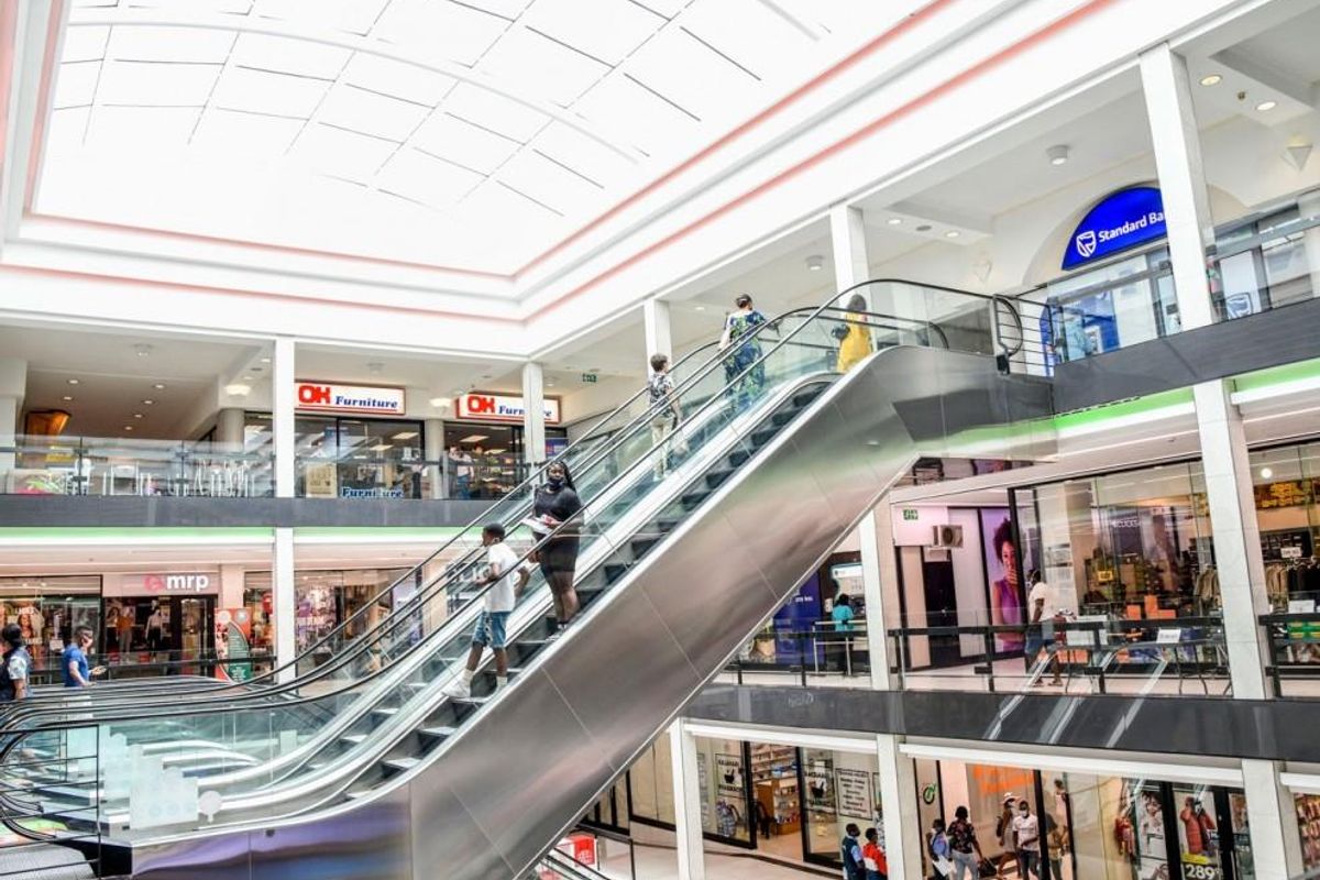 Interior view of a multi-level retail shopping center featuring a central escalator, glass railings, and a large overhead skylight.