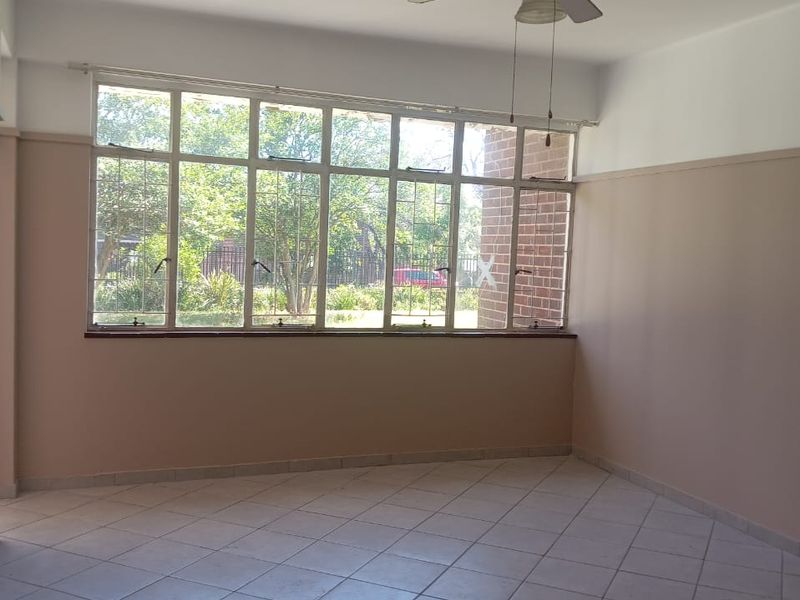 An empty living room featuring tiled flooring, a ceiling fan, and large windows providing natural light and garden views.