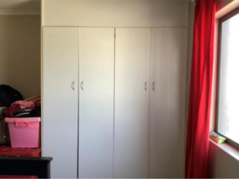 A bedroom featuring white built-in cupboards providing ample storage space, situated next to a window with red curtains.