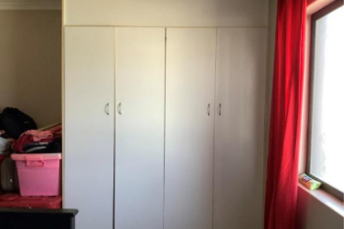 A bedroom featuring white built-in cupboards providing ample storage space, situated next to a window with red curtains.