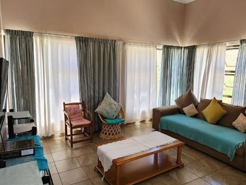 A spacious living room featuring tiled flooring, ornate ceiling cornicing, a decorative ceiling rose, and large windows allowing for ample natural light.
