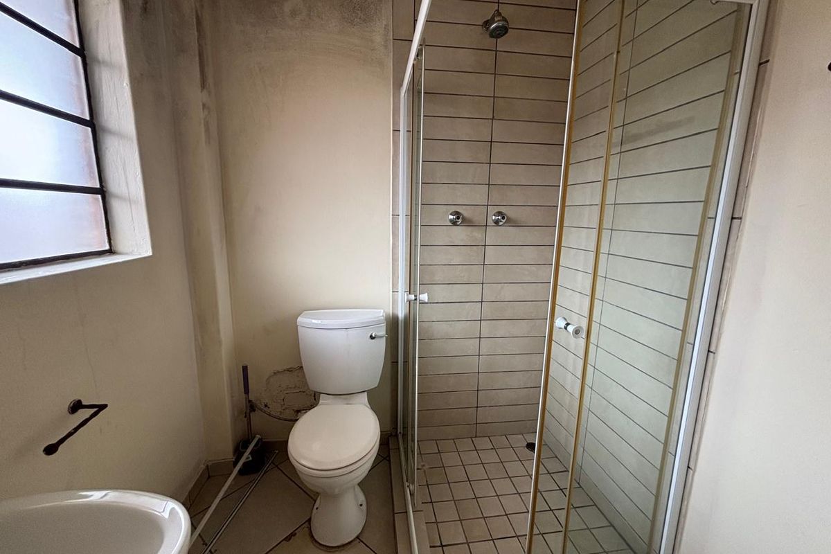 A dated bathroom featuring a glass-enclosed shower, a standard toilet, and tiled flooring, requiring significant cosmetic renovation and wall repairs.