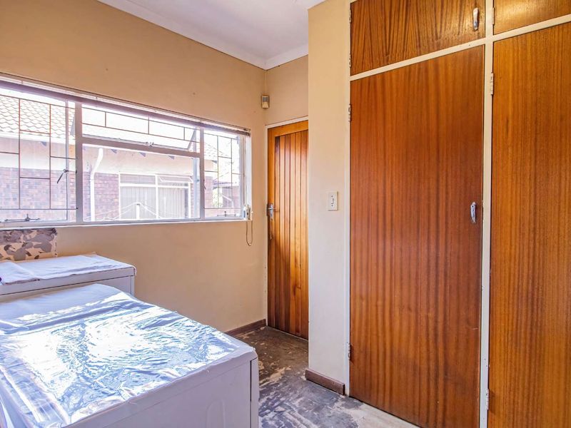 A bedroom featuring built-in wooden cupboards, a wooden door, and an aluminium window with security bars, requiring cosmetic updates to the flooring and walls.