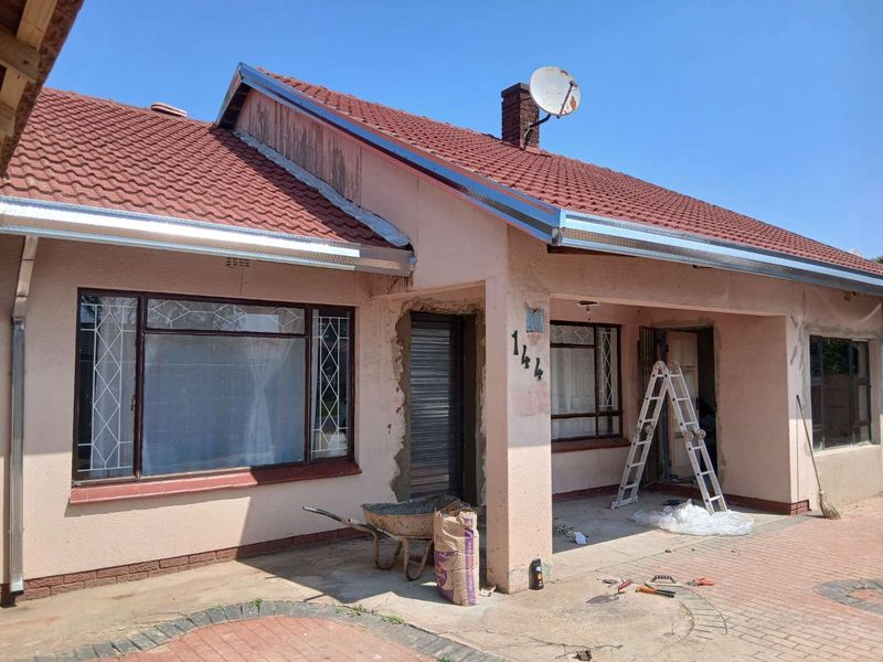Front elevation of a single-story house featuring a tiled roof, paved driveway, and ongoing exterior maintenance work around the entrance area.