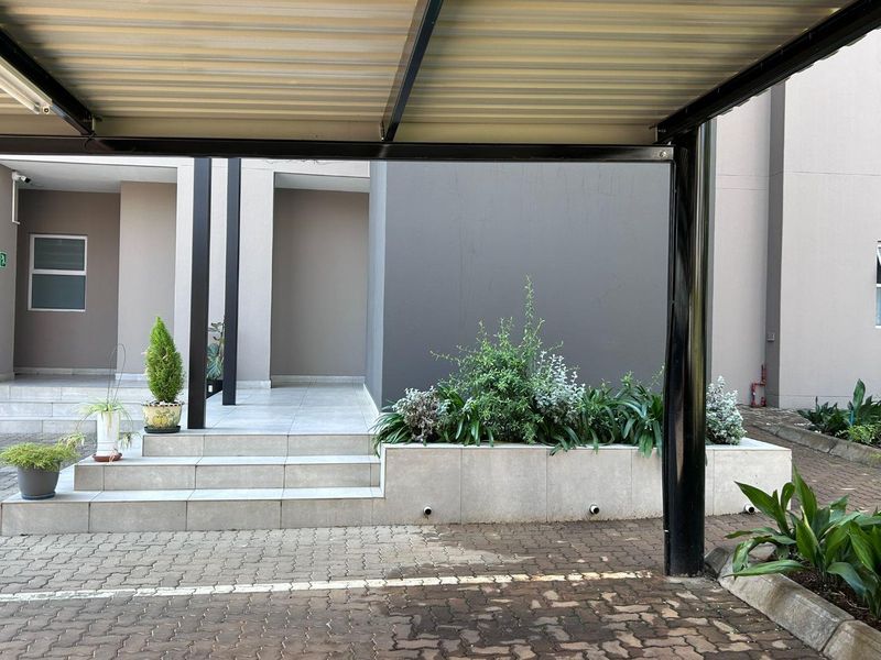 A modern apartment entrance featuring a covered carport, tiled steps, and a neat raised planter box with established landscaping.