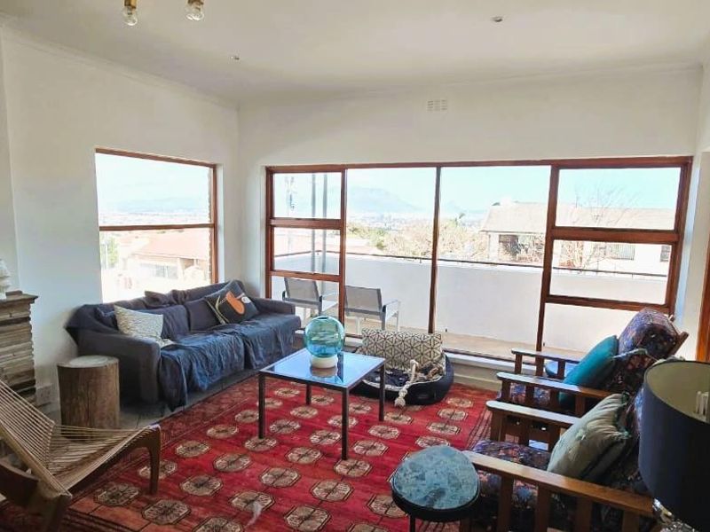 A bright living room featuring a stone fireplace, large timber-framed windows, and sliding doors that open onto an outdoor balcony with scenic views.