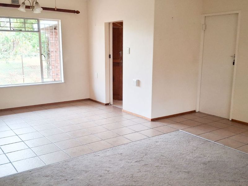 A spacious, vacant living area featuring a combination of tiled and carpeted flooring, large windows with security bars, and neutral wall finishes.
