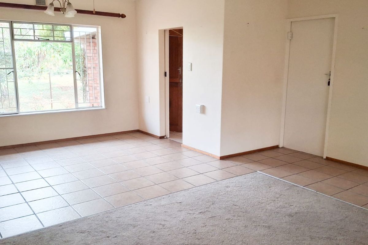 A spacious, vacant living area featuring a combination of tiled and carpeted flooring, large windows with security bars, and neutral wall finishes.