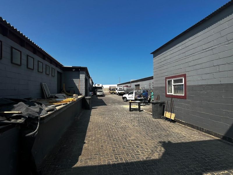 An industrial exterior rear yard featuring paved driveway access between two concrete block warehouse structures with corrugated metal roofing.
