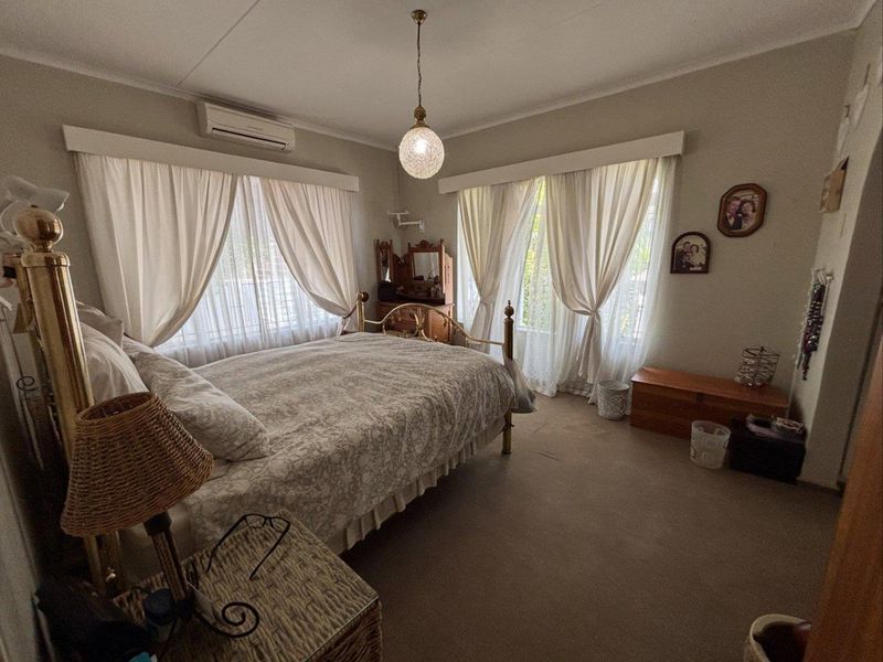 A spacious bedroom featuring neutral walls, carpeted flooring, and dual windows with curtain treatments, equipped with a wall-mounted air conditioning unit.
