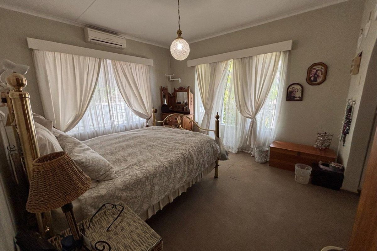 A spacious bedroom featuring neutral walls, carpeted flooring, and dual windows with curtain treatments, equipped with a wall-mounted air conditioning unit.