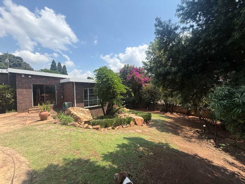 A well-maintained rear garden featuring a face brick exterior, established trees, a stone rockery, and a paved patio area accessible via sliding doors.