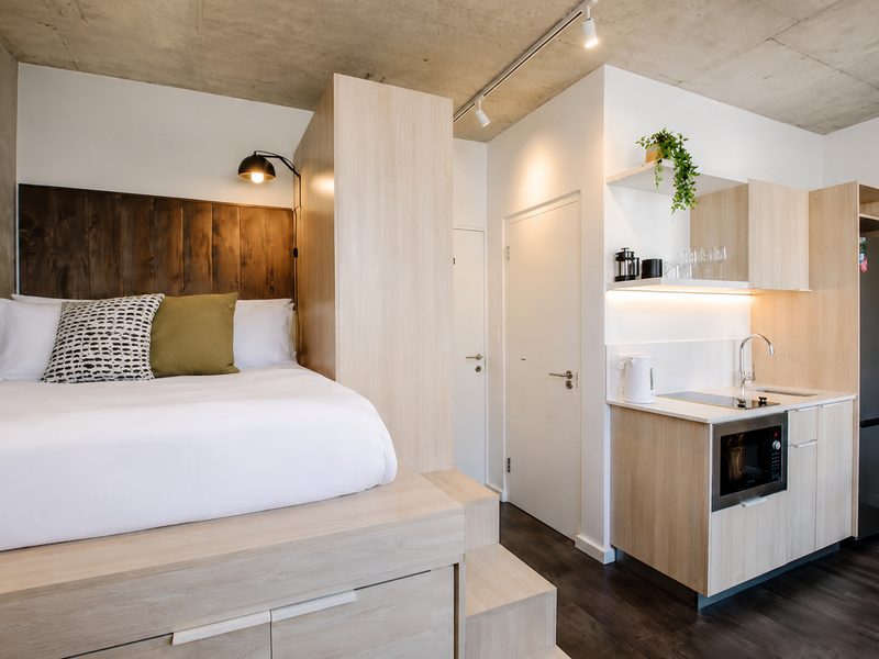 Modern open-plan studio apartment featuring an exposed concrete ceiling, custom built-in bed platform, and a compact, well-appointed kitchenette with integrated appliances.