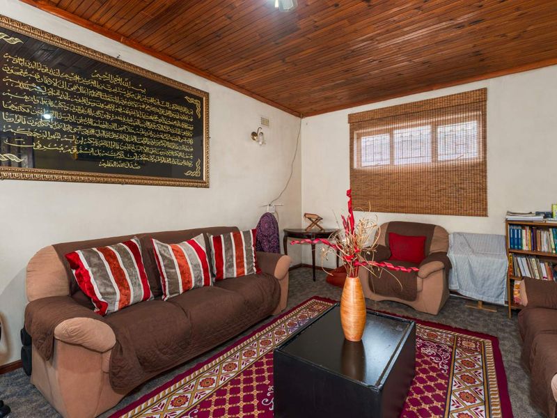 A living room featuring a timber-clad ceiling, neutral walls, and a window with a bamboo blind, offering a dated but functional living space.