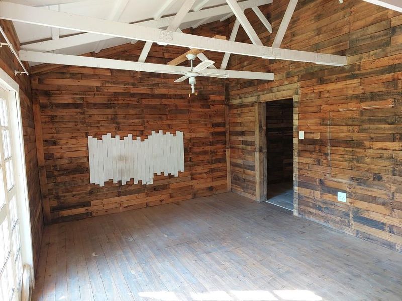 An empty living room featuring rustic timber wall cladding, exposed white-painted roof trusses, timber flooring, and a ceiling fan.