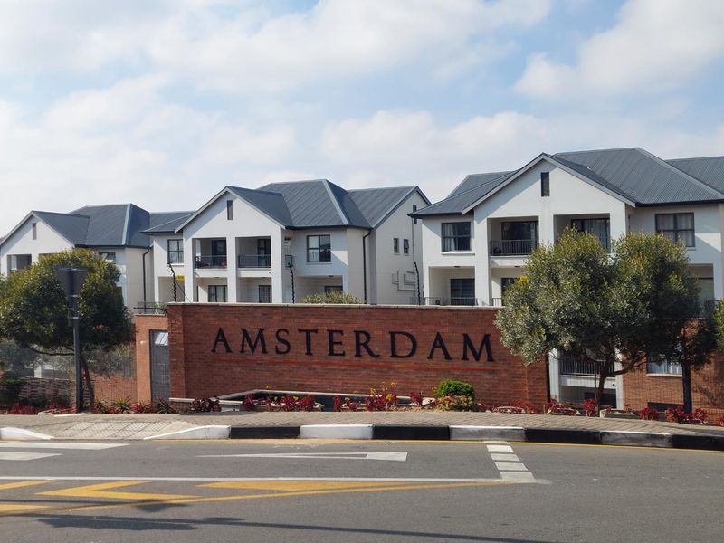 Street view of the Amsterdam apartment complex featuring a prominent brick entrance wall, multi-story residential buildings with metal roofing, and landscaped garden beds.