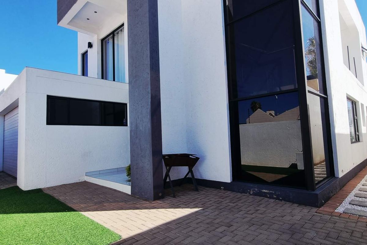 Modern exterior rear view of the property featuring paved patio area, artificial lawn, and large glass window panels.