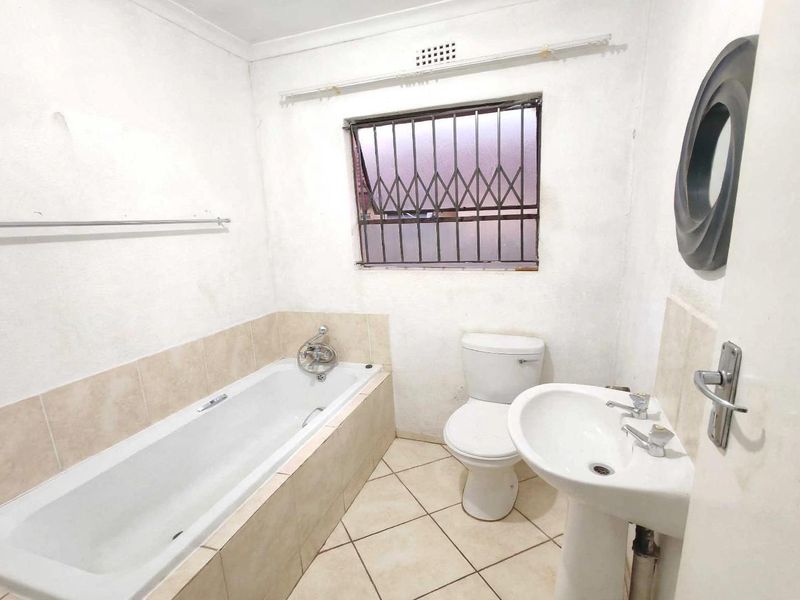 A functional bathroom featuring a bathtub, pedestal basin, and toilet, with security bars on the window and tiled finishes throughout.