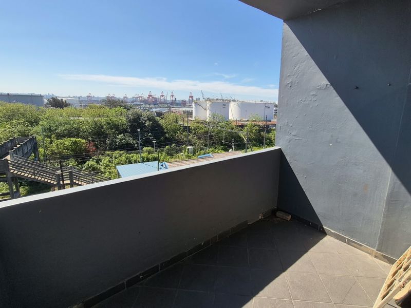 An open balcony featuring tiled flooring and a concrete balustrade, offering an elevated view of the surrounding industrial area and greenery.