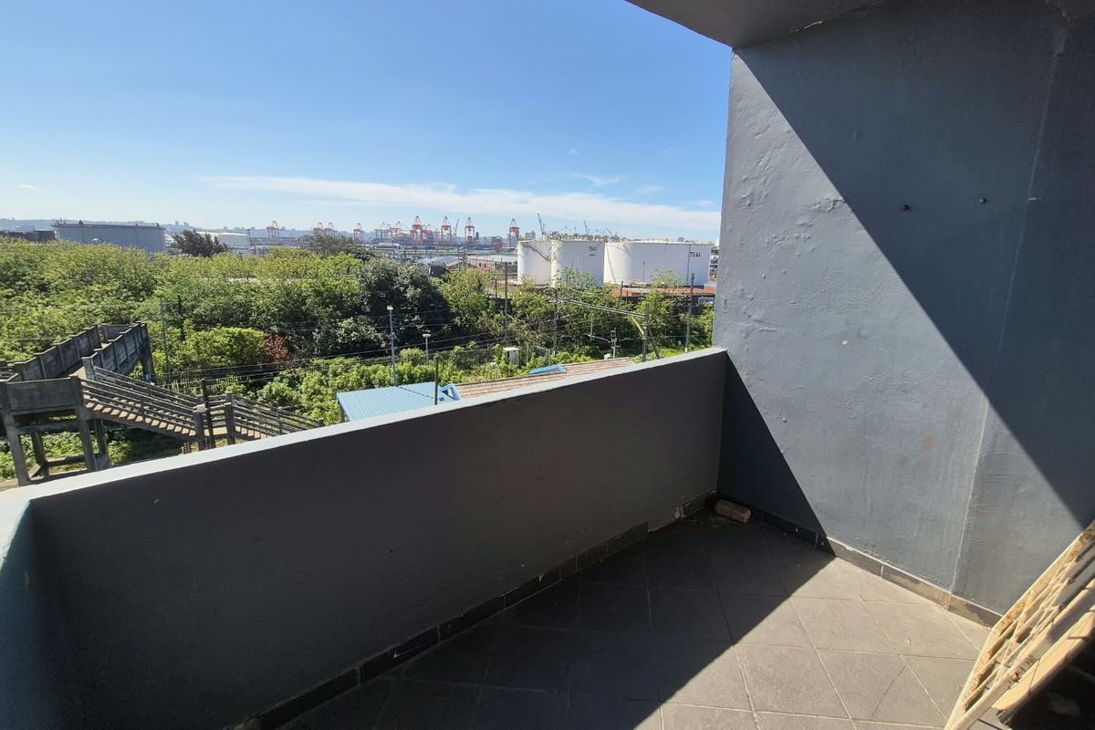 An open balcony featuring tiled flooring and a concrete balustrade, offering an elevated view of the surrounding industrial area and greenery.