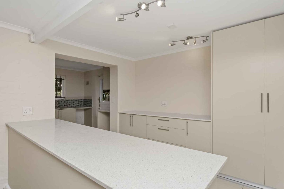 A clean, modern kitchen area featuring neutral cabinetry, stone countertops, and track lighting, with an open-plan layout connecting to an adjacent scullery or prep space.