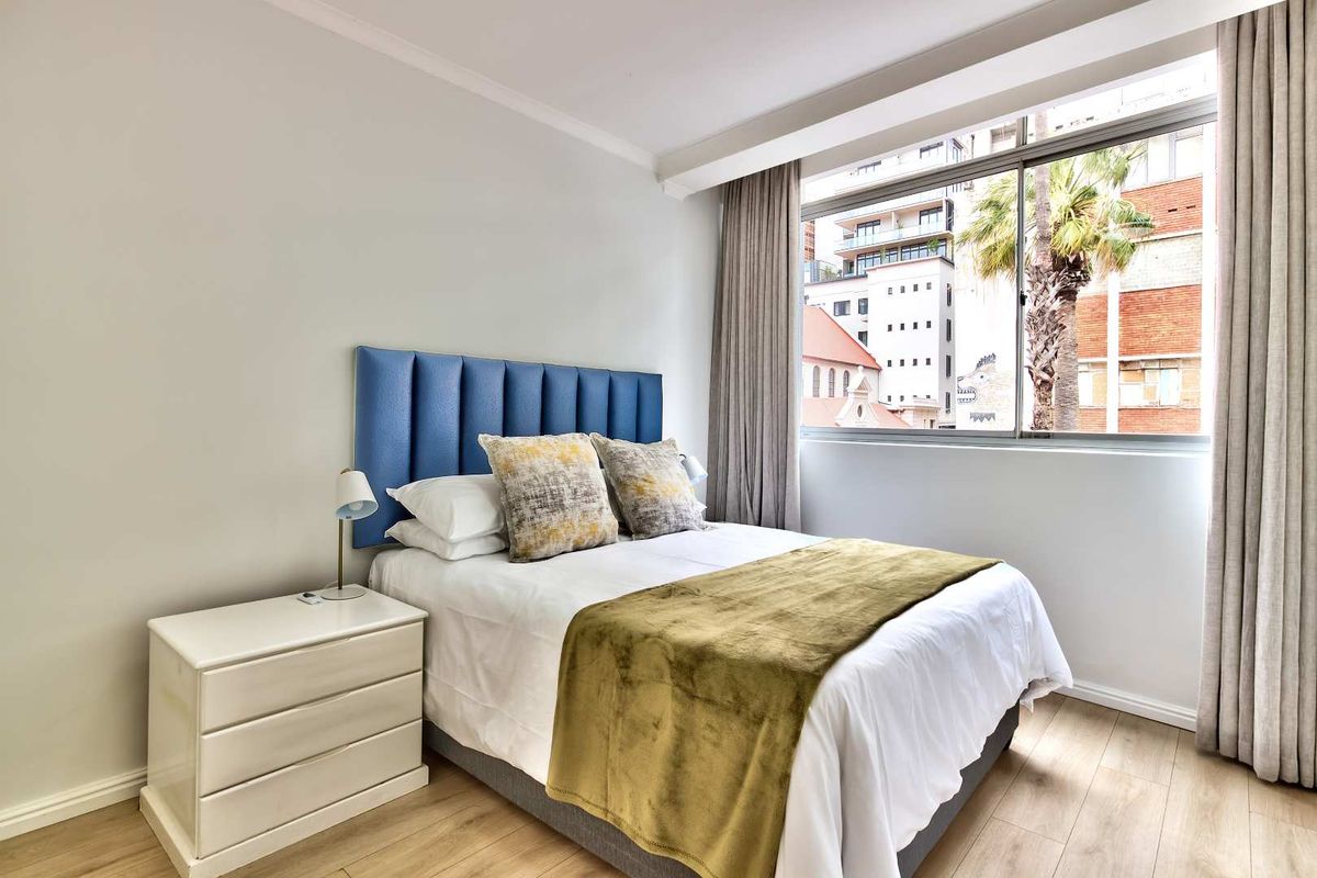 A well-maintained bedroom featuring light laminate flooring, neutral walls, and a large window providing natural light and urban views.