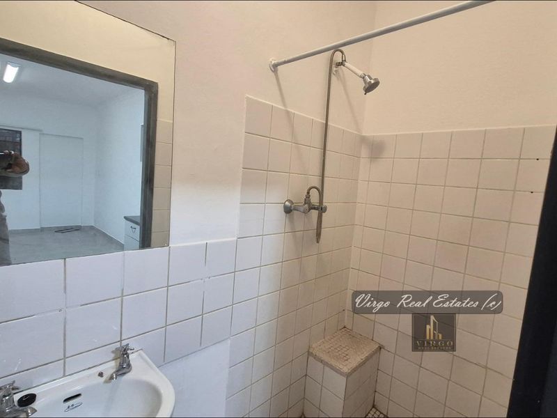 A functional bathroom featuring a pedestal washbasin, wall-mounted mirror, and a tiled shower area with exposed plumbing fixtures.