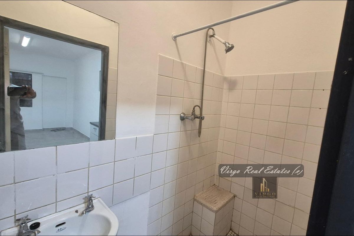 A functional bathroom featuring a pedestal washbasin, wall-mounted mirror, and a tiled shower area with exposed plumbing fixtures.