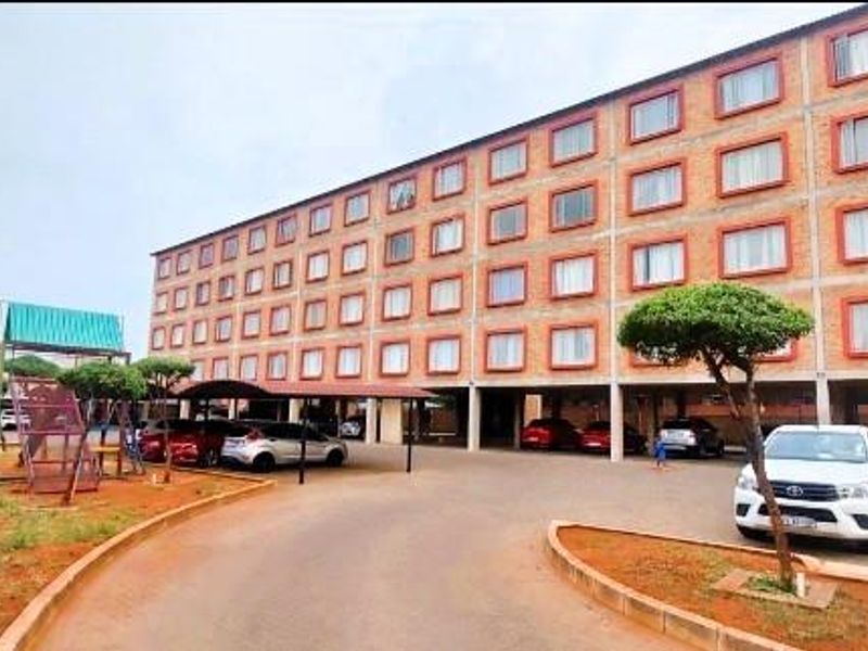 Exterior view of a multi-story apartment complex featuring a brick facade, covered parking bays, and a paved driveway with surrounding landscaped garden areas.