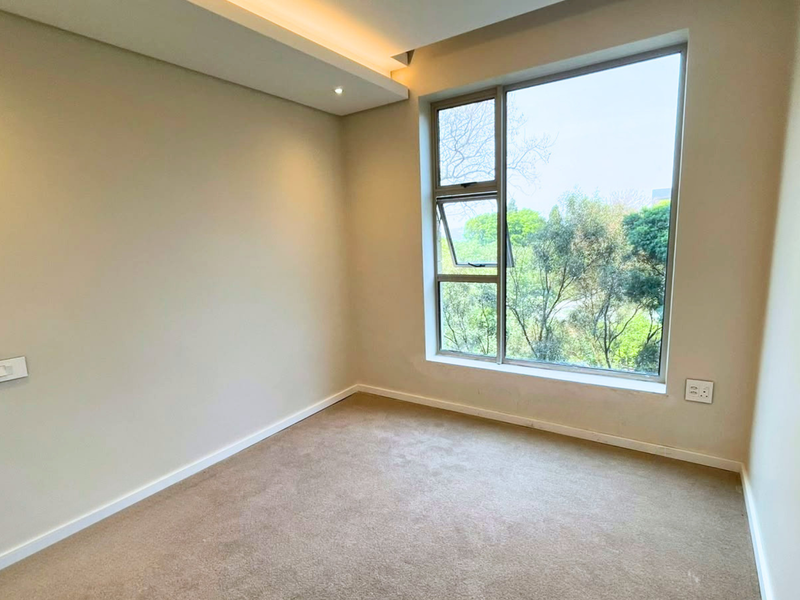 A bright, empty bedroom featuring neutral walls, carpeted flooring, recessed ceiling lighting, and a large window with views of the surrounding greenery.