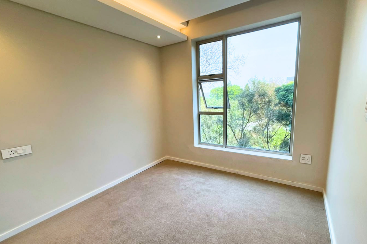 A bright, empty bedroom featuring neutral walls, carpeted flooring, recessed ceiling lighting, and a large window with views of the surrounding greenery.
