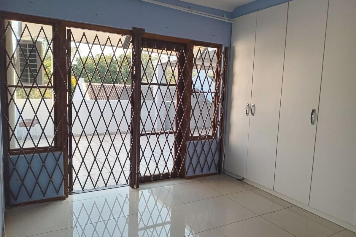 A bright, empty bedroom featuring built-in cupboards, tiled flooring, and a sliding door with a security gate leading to an outdoor area.