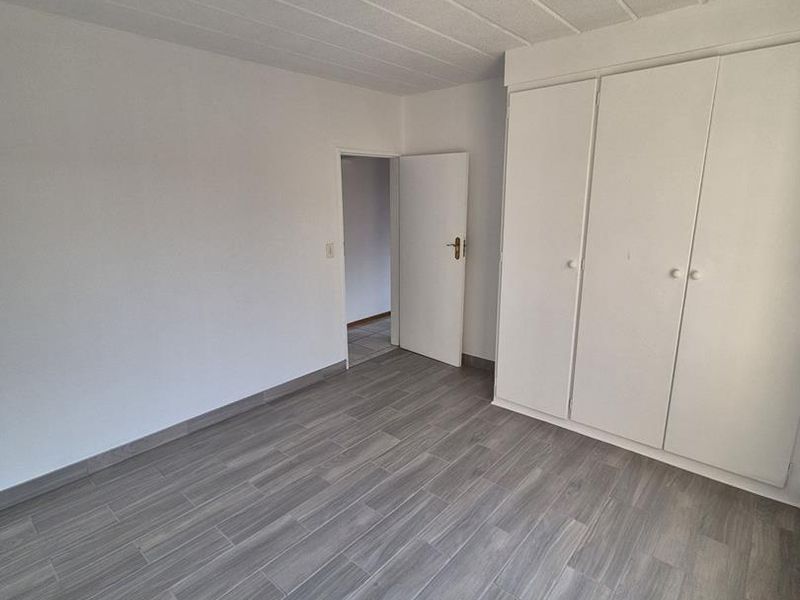 A bright, empty bedroom featuring modern grey wood-look tiled flooring and white built-in cupboards, offering a clean and neutral space.
